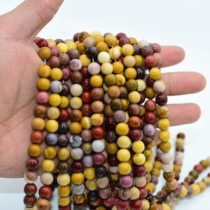 May include: A strand of round, multi-colored gemstone beads. The beads are a mix of yellow, brown, red, and purple. The beads are strung on a clear elastic cord.