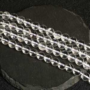 May include: A strand of clear quartz beads, each bead is round and smooth. The beads are arranged in a line on a dark grey surface.