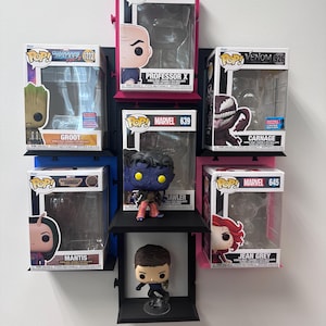 May include: A wall-mounted display unit with several Funko Pop! figures. The figures include Groot, Professor X, Carnage, Mantis, Jean Grey, and others. The display has black, blue, and pink shelves.