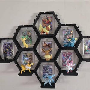 May include: A black hexagonal wall-mounted display shelf system featuring multiple compartments. Each section holds a clear protective case containing a trading card and a small plastic figurine of a creature, including Eevee and its evolutions.