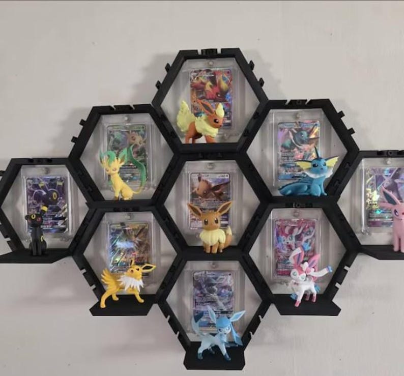May include: A black hexagonal wall-mounted display shelf system featuring multiple compartments. Each section holds a clear protective case containing a trading card and a small plastic figurine of a creature, including Eevee and its evolutions.