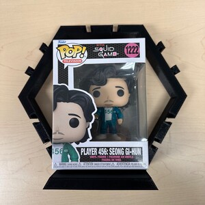 May include: A Funko Pop Television vinyl figure of Player 456: Seong Gi-hun from Squid Game, number 1222, inside its original window box packaging. The figure is displayed in front of a black, hexagonal, 3D-printed plastic display stand.