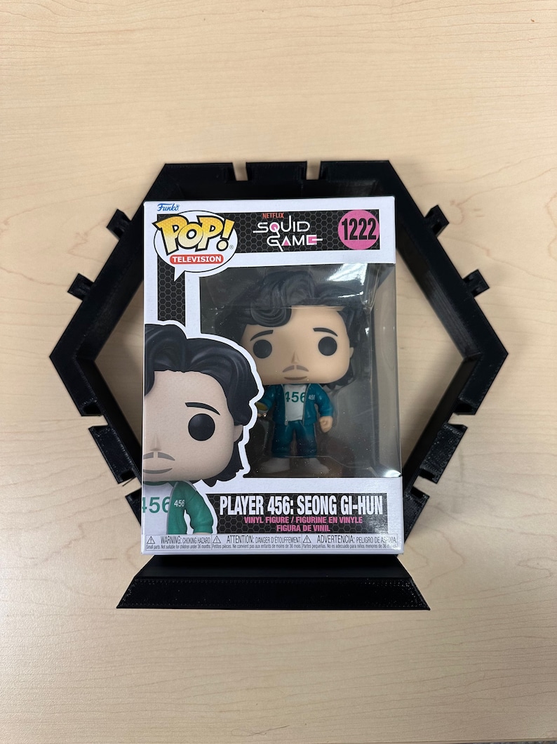 May include: A Funko Pop Television vinyl figure of Player 456: Seong Gi-hun from Squid Game, number 1222, inside its original window box packaging. The figure is displayed in front of a black, hexagonal, 3D-printed plastic display stand.