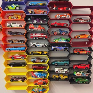 May include: A wall-mounted display system featuring interconnected hexagonal shelves in purple, orange, yellow, black, and red. The shelves hold a collection of various die-cast toy cars, including sports cars, a fire truck, and a dinosaur-shaped vehicle.