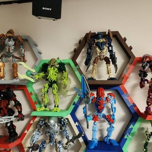 May include: A collection of Bionicle action figures displayed on a wall-mounted honeycomb shelving system. The figures feature various colors including orange, lime green, dark blue, red, and silver, with articulated limbs and weapons.