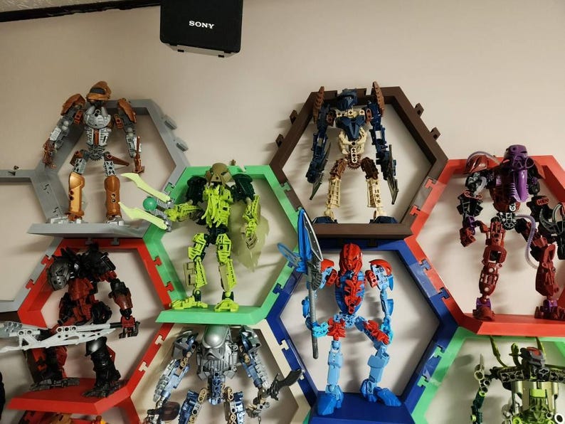 May include: A collection of Bionicle action figures displayed on a wall-mounted honeycomb shelving system. The figures feature various colors including orange, lime green, dark blue, red, and silver, with articulated limbs and weapons.