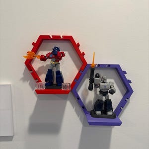 K&ouml;nnte beinhalten: Zwei sechseckige Wandregale, eines in Rot und eines in Lila, die jeweils eine Kunststoff-Actionfigur halten. Das rote Regal enth&auml;lt einen blau-roten Roboter mit einer orangefarbenen Axt, w&auml;hrend das lila Regal einen grauen Roboter mit Schwert zeigt.