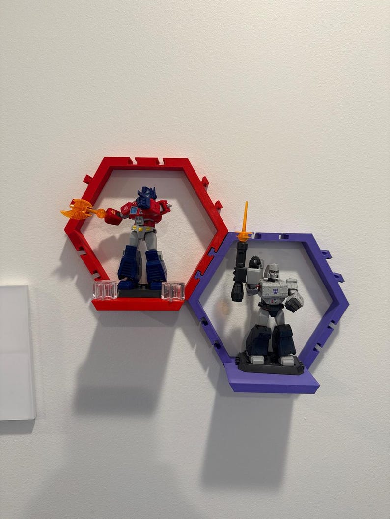 K&ouml;nnte beinhalten: Zwei sechseckige Wandregale, eines in Rot und eines in Lila, die jeweils eine Kunststoff-Actionfigur halten. Das rote Regal enth&auml;lt einen blau-roten Roboter mit einer orangefarbenen Axt, w&auml;hrend das lila Regal einen grauen Roboter mit Schwert zeigt.