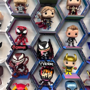 K&ouml;nnte beinhalten: Eine Sammlung von Vinylfiguren auf einem modularen, sechseckigen Wandregalsystem. Die Figuren zeigen verschiedene Charaktere mit gro&szlig;en K&ouml;pfen und stilisierten Merkmalen, einige stehen auf kleinen Podesten mit Beschriftungen wie Carnage und Venom.
