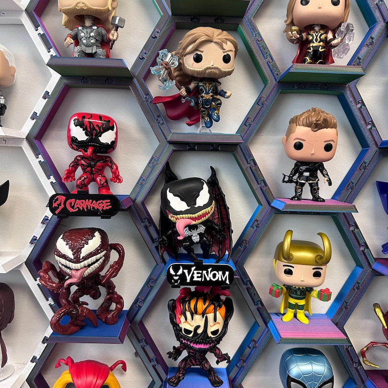 K&ouml;nnte beinhalten: Eine Sammlung von Vinylfiguren auf einem modularen, sechseckigen Wandregalsystem. Die Figuren zeigen verschiedene Charaktere mit gro&szlig;en K&ouml;pfen und stilisierten Merkmalen, einige stehen auf kleinen Podesten mit Beschriftungen wie Carnage und Venom.