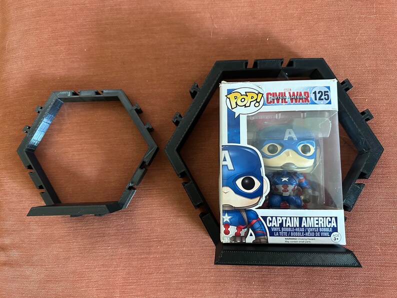 May include: Two black hexagonal display frames made of plastic, one empty and one containing a Captain America Funko Pop vinyl bobble-head figure in its original box, labeled Civil War 125, featuring the character in a blue and red suit.
