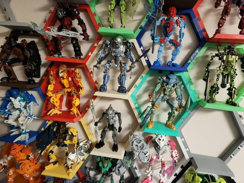 May include: A collection of Bionicle action figures displayed on a wall-mounted honeycomb shelving system. The figures feature various colors including red, blue, green, yellow, and silver, each posed on individual hexagonal shelves.