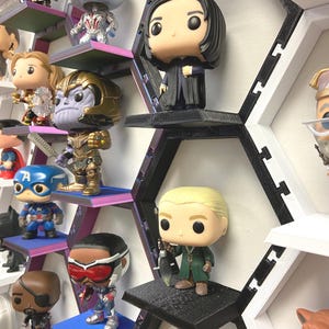 K&ouml;nnte beinhalten: Eine Sammlung von Vinyl-Wackelkopf-Figuren, die auf einem modularen, schwarz-wei&szlig;en, wabenf&ouml;rmigen Wandregalsystem ausgestellt sind. Die Figuren zeigen verschiedene Charaktere mit gro&szlig;en K&ouml;pfen und kleinen K&ouml;rpern auf einzelnen Regalb&ouml;den.