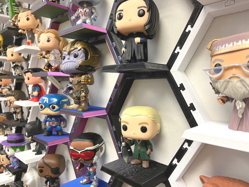 K&ouml;nnte beinhalten: Eine Sammlung von Vinyl-Wackelkopf-Figuren, die auf einem modularen, schwarz-wei&szlig;en, wabenf&ouml;rmigen Wandregalsystem ausgestellt sind. Die Figuren zeigen verschiedene Charaktere mit gro&szlig;en K&ouml;pfen und kleinen K&ouml;rpern auf einzelnen Regalb&ouml;den.