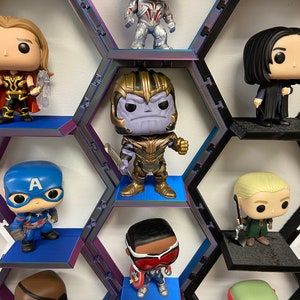 K&ouml;nnte beinhalten: Eine Sammlung von Vinylfiguren auf einem modularen Waben-Wandregalsystem. Die Figuren zeigen Charaktere mit markanten Merkmalen wie einen violetth&auml;utigen gepanzerten Titanen, einen blonden Helden mit rotem Umhang und einen blau maskierten Helden.