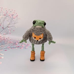May include: A green knitted frog wearing a gray sweater with an orange fox design and brown boots.