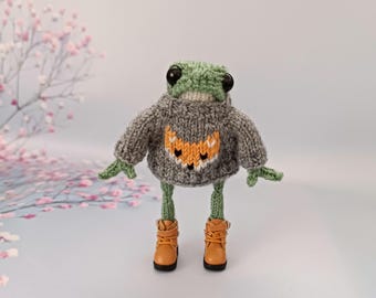 May include: A green knitted frog wearing a gray sweater with an orange fox design and brown boots.