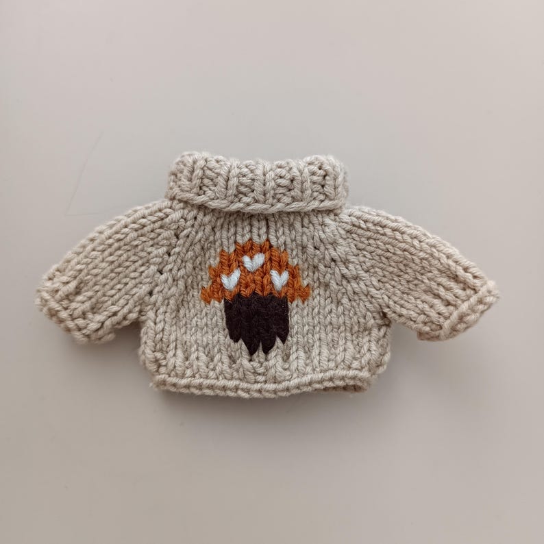 May include: A beige knitted sweater with a brown and orange mushroom design. The mushroom has white hearts on top.