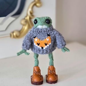 May include: A green knitted frog wearing a gray sweater with an orange and white pattern and brown leather boots.