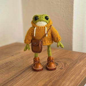 May include: A knitted frog figure wearing a mustard yellow hoodie, brown leather boots, and a matching crossbody bag. The frog has green and white accents and black eyes. The frog is standing on a wooden surface.