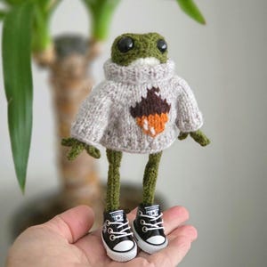 Könnte beinhalten: Handgefertigte grüne Froschfigur mit grauem Pullover mit Eicheldesign und schwarzen Converse-Sneakern. Der Frosch hat große schwarze Augen und grüne Gliedmaßen. Ein skurriles, handgefertigtes Objekt.