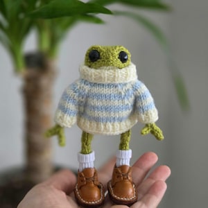 May include: A handmade crochet frog doll wearing a blue and white striped sweater, white socks, and brown leather shoes. The frog has large black eyes and green limbs. The doll is a whimsical, handcrafted item.