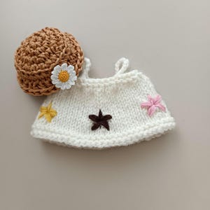 May include: A white crocheted doll dress with brown, pink, and yellow embroidered flowers and stars. The dress is paired with a brown crocheted hat.
