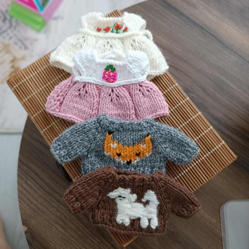 May include: Four knitted doll clothes, including a white dress with red flowers, a pink dress with a strawberry, a gray sweater with a fox, and a brown sweater with a white dog.