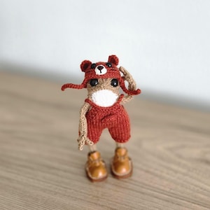 Peut inclure: Poupée au crochet faite à la main, vêtue d'un costume d'ours. La poupée porte un chapeau d'ours rouge, une salopette et des chaussures marron. Le corps de la poupée est beige et blanc, avec des yeux noirs. La poupée est sur une surface en bois.