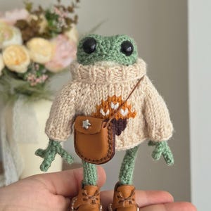 Könnte beinhalten: Eine gehäkelte grüne Froschpuppe, die einen beigen Strickpullover mit einem braunen Eichelmotiv auf der Vorderseite trägt. Der Frosch hat schwarze Kunststoffaugen, eine kleine braune Umhängetasche aus Kunstleder mit silbernem Stern und braune Stiefel.