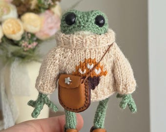 Puede incluir: Una muñeca de rana verde tejida a ganchillo que lleva un suéter de punto beige con un diseño de bellota marrón en la parte delantera. La rana tiene ojos de plástico negro, un pequeño bolso bandolera de piel sintética marrón con una estrella.