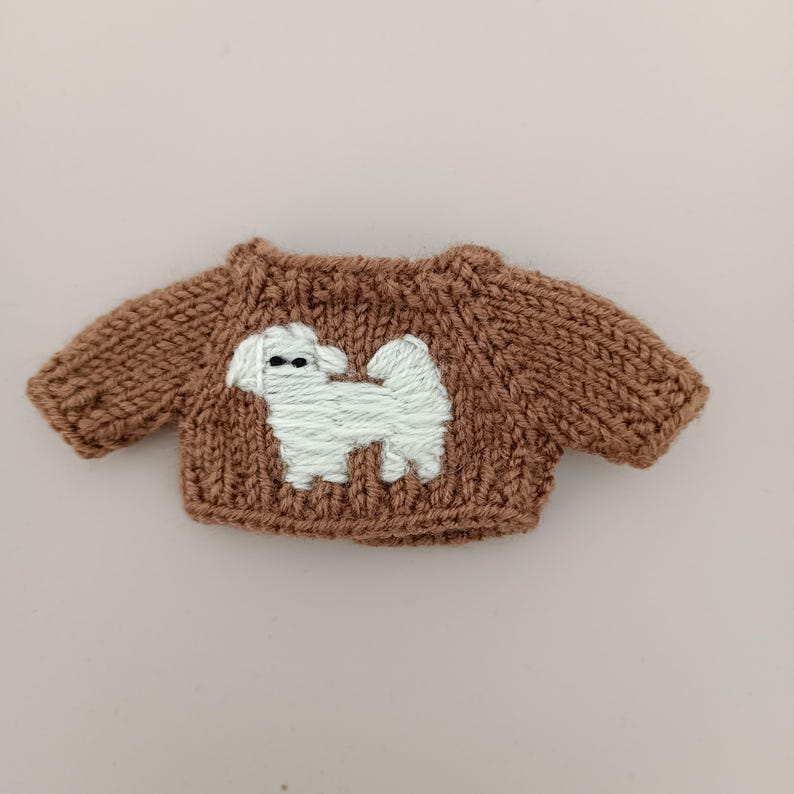 May include: A brown knitted sweater with a white embroidered dog design. The sweater is small and appears to be for a doll or small toy.