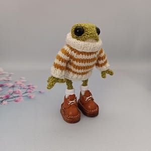 May include: A green crocheted frog wearing a white and brown striped sweater and brown shoes.