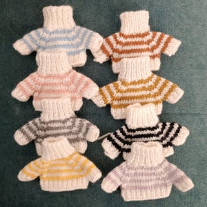 May include: Eight miniature knitted sweaters in various colors and striped patterns. The sweaters are white with stripes of blue, brown, pink, yellow, gray, black, and purple.