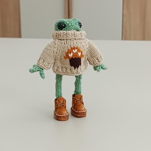 Op de afbeelding: Een groene gebreide kikker die een beige trui draagt met een bruin paddenstoelontwerp en bruine leren laarzen.