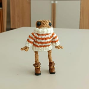 May include: A brown crocheted frog wearing a white and orange striped sweater and brown boots.