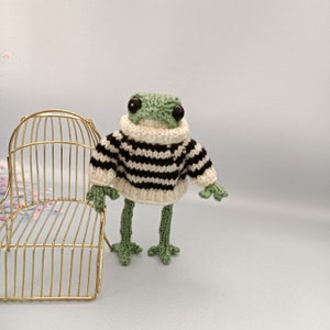 Könnte beinhalten: Ein grüner gehäkelter Frosch, der einen schwarz-weiß gestreiften Pullover trägt. Der Frosch steht auf einem goldenen Drahtstuhl.