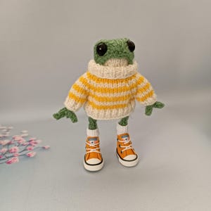 May include: A green crocheted frog wearing a yellow and white striped sweater and orange sneakers.