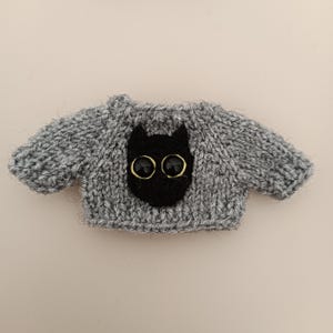 May include: A gray knitted sweater with a black cat face appliqu&eacute; and gold button eyes.