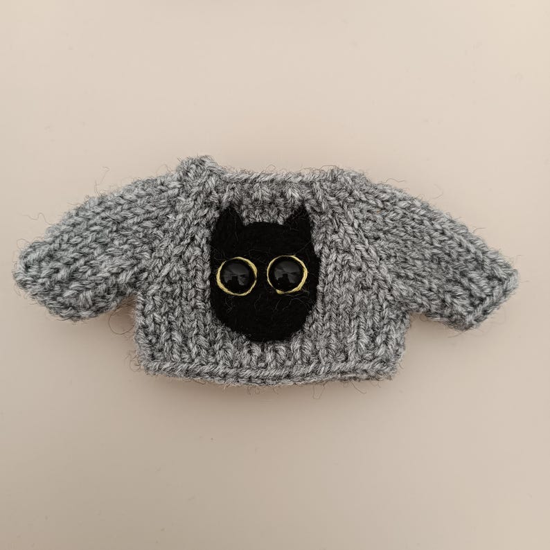 May include: A gray knitted sweater with a black cat face appliqu&eacute; and gold button eyes.
