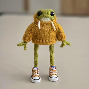 Könnte beinhalten: Ein grüner gestrickter Frosch, der einen gelben gestrickten Hoodie und braune und weiße Turnschuhe trägt.