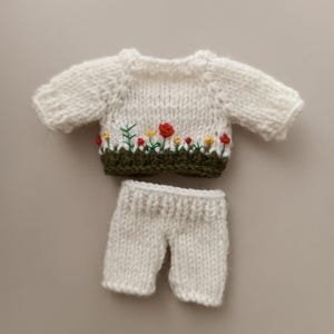 May include: A white knitted sweater with a green and red embroidered flower design. The sweater is paired with white knitted pants.