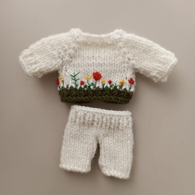 May include: A white knitted sweater with a green and red embroidered flower design. The sweater is paired with white knitted pants.