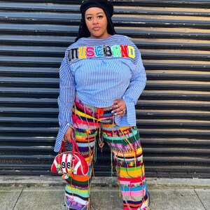 May include: A woman wearing a blue and white striped shirt with the word "MUSEBRAND" across the chest. She is also wearing colorful crochet pants, a black beret, and carrying a red handbag with the number "96" on it. The outfit is a mix of textures and colors.