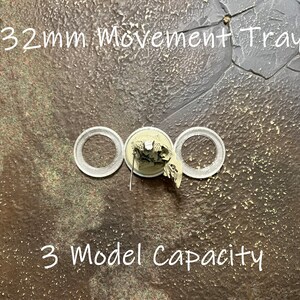May include: A 32mm movement tray with three circular cutouts for miniature figures. The tray is made of a brown material with a textured surface. The text "32mm Movement Tray" and "3 Model Capacity" is printed on the tray.