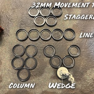 May include: A set of 32mm movement trays for tabletop games. The trays are arranged in different formations, including a line, a column, a wedge, and a staggered pattern. The trays are made of black plastic and have a smooth, circular shape.