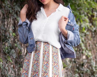 May include: A woman wearing a white button-down shirt, a denim jacket, and a colorful patterned skirt. The skirt has vertical stripes and a floral design.