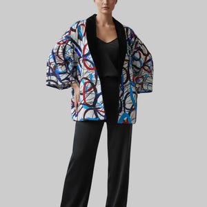 Puede incluir: Chaqueta estampada estilo kimono con cuello y puños de terciopelo negro. La chaqueta presenta un fondo blanco con diseños abstractos azules y rojos. La modelo lleva un top negro y pantalones negros de pernera ancha.