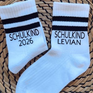 Könnte beinhalten: Paar weiße Socken mit schwarzen Streifen am oberen Rand. Eine Socke trägt den Aufdruck "SCHULKIND 2026", die andere "SCHULKIND LEVIAN" in Schwarz. Die Socken liegen auf einer gewebten, braunen Oberfläche.