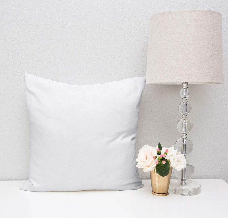 May include: A white pillow sits on a white surface next to a lamp with a beige lampshade and a gold vase with white flowers.